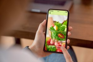 A person is holding a smartphone displaying a vibrant image of a fresh salad with cherry tomatoes and greens, accompanied by a prominent "Order Now" button. The background includes a blurred view of a desk with a laptop. This image emphasizes the importance of high-quality visual content for a standout food truck design website.