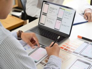 Designer sketching mobile layouts on paper, with a laptop showing wireframes, focused on user experience. Image represents the best podcast website design.