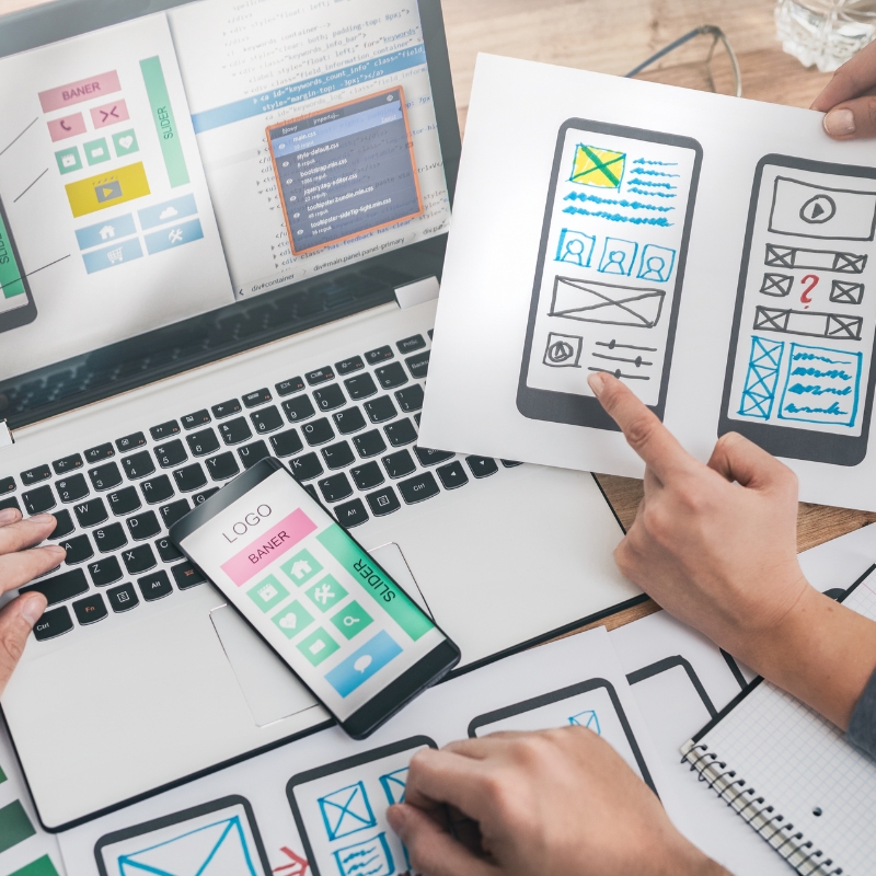 A team at a website design agency reviewing mobile UI wireframes, a phone prototype, and laptop code.